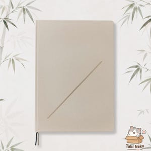 Può includere: Un taccuino rettangolare con copertina color beige chiaro tinta unita, caratterizzato da una singola linea diagonale in rilievo sulla parte anteriore. Un piccolo segnalibro a nastro nero sporge dal bordo inferiore.