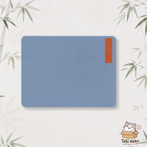 May include: A rectangular, solid slate blue mouse pad with rounded corners. A small, vertical, orange rectangular tag is attached to the right side, featuring the text "Tabi Neko" in a simple, dark font. The surface is smooth and matte.