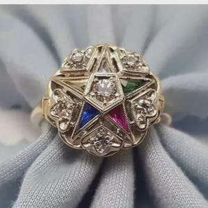 May include: A gold ring featuring a central five-pointed star emblem. The star is adorned with a clear center stone, a blue stone, a red stone, and a green stone. The surrounding metalwork includes multiple clear stones set in a star-shaped frame.
