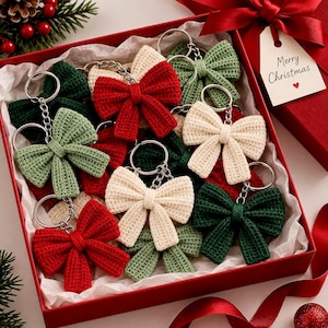 May include: A collection of crochet bow keychains in red, cream, and green colours, each attached to a silver metal keyring. The keychains are arranged inside a red gift box lined with white tissue paper, featuring a tag that reads Merry Christmas.