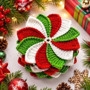 May include: A circular crochet piece featuring a radial pattern of overlapping petal-like segments in alternating red, white, and green yarn. The textured, hand-stitched design radiates from a central white ring, creating a layered, festive appearance.
