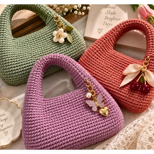 Può includere: Tre borse all'uncinetto nei colori verde salvia, rosa corallo e lavanda. Ogni borsa presenta un robusto manico integrato e un ciondolo in metallo dorato. La borsa verde ha un fiore, quella rosa ciliegie con un nastro, e quella lavanda una farfalla e un cuore.