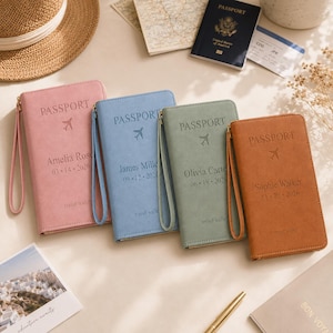 May include: Four rectangular travel wallets in pink, blue, sage green, and tan, each featuring a wrist strap, the word PASSPORT, an airplane icon, a name, and a date embossed on the front. The wallets are arranged side-by-side on a flat surface.