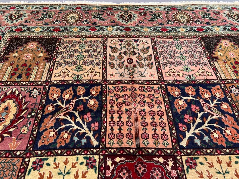 May include: A rectangular area rug featuring a grid of square panels, each decorated with intricate floral and tree motifs in shades of pink, navy blue, cream, and brown. The rug includes a wide, detailed border with repeating patterns and fringe.