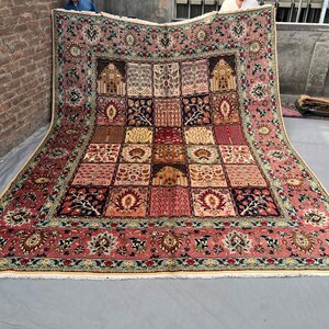 May include: A large rectangular area rug featuring a complex grid pattern of various floral and architectural motifs. The rug is primarily colored in shades of dusty pink, deep red, navy blue, and cream, with a wide decorative border and fringe.