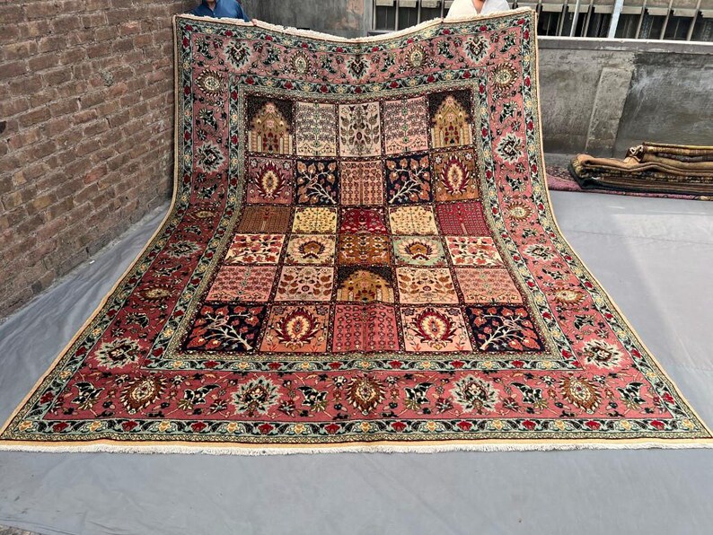 May include: A large rectangular area rug featuring a complex grid pattern of various floral and architectural motifs. The rug is primarily colored in shades of dusty pink, deep red, navy blue, and cream, with a wide decorative border and fringe.