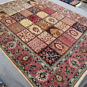 May include: A rectangular area rug featuring a patchwork design of various floral and botanical motifs in shades of pink, cream, deep red, and navy blue. The rug has a wide, ornate border with intricate patterns and a fringe along the edges.