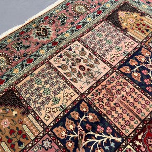 May include: A rectangular area rug featuring a grid of square panels, each decorated with intricate floral and tree motifs in shades of pink, navy blue, cream, and deep red. The rug has a wide, ornate border and white fringe along the edge.