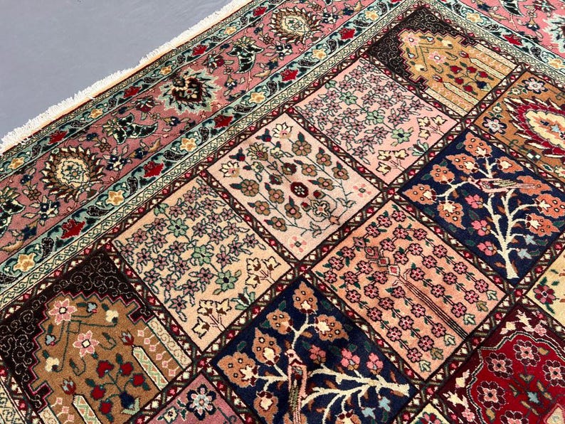 May include: A rectangular area rug featuring a grid of square panels, each decorated with intricate floral and tree motifs in shades of pink, navy blue, cream, and deep red. The rug has a wide, ornate border and white fringe along the edge.