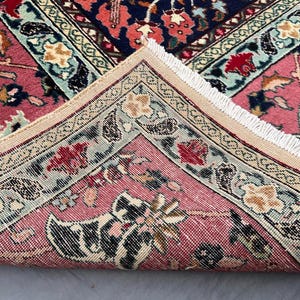 May include: A woven rug featuring a folded corner that reveals a pink background with intricate floral and paisley patterns in shades of red, navy blue, cream, and light green. The edge of the rug is finished with a short, white fringe trim.