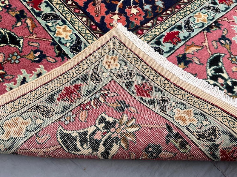May include: A woven rug featuring a folded corner that reveals a pink background with intricate floral and paisley patterns in shades of red, navy blue, cream, and light green. The edge of the rug is finished with a short, white fringe trim.