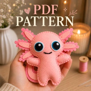May include: A pink felt axolotl plush toy with large black and blue eyes and a stitched mouth. The creature features scalloped external gills on both sides of its head. The text PDF PATTERN is displayed in white above the plush toy.
