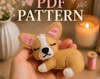Op de afbeelding: Een klein, handgemaakt vilt corgi-hondornament in beige en wit met roze binnenoren en geborduurde gesloten ogen. De tekst PDF PATTERN staat boven de figuur, die een witte vlek op het gezicht, een zwarte neus en witte pootjes heeft.
