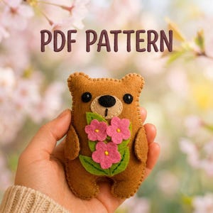 May include: A brown felt bear plush toy featuring a tan muzzle, black eyes, and a bouquet of three pink flowers with green leaves stitched to its chest. The text PDF PATTERN is displayed in brown letters above the bear against a floral background.