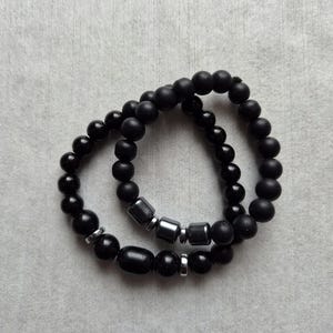 May include: Two stretch bracelets featuring matte and polished black beads. One bracelet includes three metallic cylindrical beads, while the other features smaller silver-toned spacers and a single larger oval black bead interspersed among the round beads.