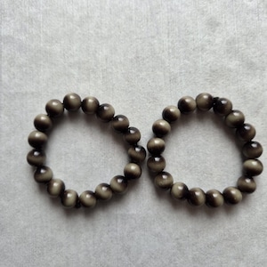 Könnte beinhalten: Zwei kreisförmige Armbänder aus glatten, kugelförmigen Perlen. Jede Perle weist ein Farbverlaufsmuster auf, das von einer dunklen, bräunlich-grauen Mitte zu helleren, cremefarbenen Rändern übergeht und einen reflektierenden Katzenaugen-Effekt erzeugt.