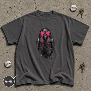 以下が含まれることがあります： チャコールグレーの半袖Tシャツで、中央には鮮やかなピンクのハート型の目からペンキが垂れているような黒いカラスのグラフィックが描かれています。クルーネックでゆったりとしたフィット感のシャツで、2本の鍵と2つのピンバッジが添えられています。