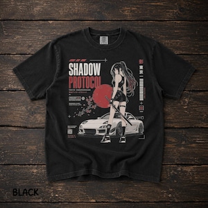 Op de afbeelding: Een zwart t-shirt met een grafische print van een personage met een zwaard voor een witte sportwagen en een rode zon. De tekst luidt SHADOW PROTOCOL, TOKYO UNDERGROUND, en bevat Japanse karakters en technische ontwerpelementen.