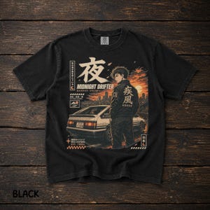 Puede incluir: Una camiseta negra de manga corta con un estampado gráfico de una persona de pie junto a un coche deportivo blanco al atardecer. El diseño incluye texto en japonés, las palabras MIDNIGHT DRIFTER y un fondo de horizonte urbano en tonos naranja.