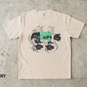 May include: An ivory short-sleeve t-shirt featuring a black ink graphic of two overlapping faces with dark, smudged eye makeup. A bright mint green paint-like splash is printed across the centre of the faces. The word IVORY is printed in black.