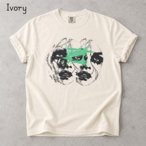 Könnte beinhalten: Ein elfenbeinfarbenes Kurzarm-T-Shirt mit einem schwarz-weißen Grafikdruck von zwei überlappenden Gesichtern und einer hellgrünen abstrakten Form über den Augen. Das T-Shirt hat einen Rundhalsausschnitt und ein Etikett am Kragen mit dem Text M-M-M.