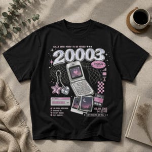 Puede incluir: Una camiseta negra con un estampado gráfico que incluye el texto "CALLS WERE MEANT TO BE MISSED" y "20003". El diseño presenta un teléfono plegable plateado, un dije de estrella y corazón, una barra de carga e iconos digitales.
