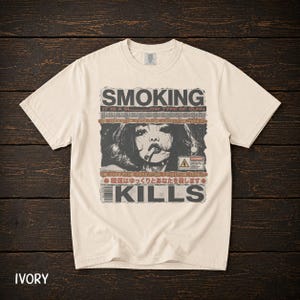 Puede incluir: Una camiseta de manga corta color marfil con un estampado gráfico de una persona fumando. El texto dice SMOKING IT IS A SLOW TYPE OF BURN y KILLS, junto con caracteres japoneses y un símbolo de advertencia en un diseño tipo periódico.