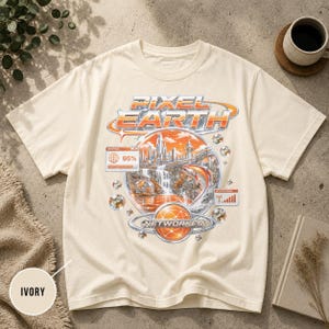 Könnte beinhalten: Ein elfenbeinfarbenes Kurzarm-T-Shirt mit einem Grafikdruck mit dem Text PIXEL EARTH und NETWORKED. Das Design zeigt eine futuristische Stadt, einen Wasserfall, einen springenden Fisch, einen Ladebalken bei 95% und ein Signalstärkesymbol in Orange und Silber.