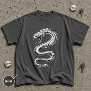 May include: A charcoal gray short-sleeve t-shirt featuring a large, metallic silver graphic of a coiled, aggressive snake with sharp fangs and detailed scales centered on the chest. The shirt has a crew neckline and a standard, relaxed fit.