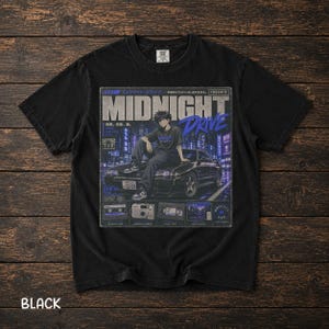Könnte beinhalten: Ein schwarzes Kurzarm-T-Shirt mit einem Grafikdruck mit der Aufschrift MIDNIGHT DRIVE. Das Design zeigt eine Anime-Figur, die auf einem schwarzen Sportwagen vor einer Stadtkulisse sitzt, ergänzt durch kleine Abbildungen einer Kassette und Kamera.