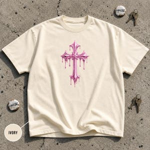 May include: An ivory short-sleeve t-shirt featuring a metallic pink cross graphic centered on the chest. The cross design has a liquid, dripping effect. The shirt includes a small circular label with the text IVORY and Gorillaz branding.