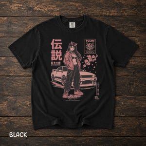 May include: A black short-sleeve t-shirt featuring a pink graphic print of a character with horns standing in front of a sports car. The design includes cherry blossom branches, Japanese text, and the word YUJIN in a rectangular frame.