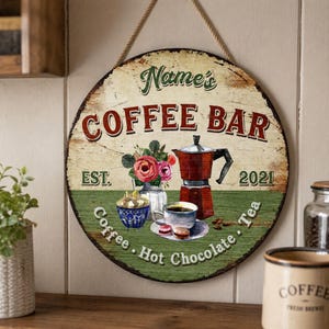May include: A round wooden sign with a distressed cream and green finish, featuring the text "Name's COFFEE BAR" and "EST. 2021". The graphic includes a red moka pot, a cup of coffee, a bowl of sugar, and flowers. The text "Coffee, Hot Chocolate, Tea" is below.