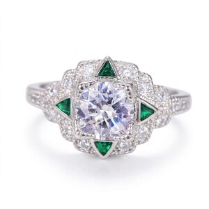 May include: A silver-toned ring featuring a large, round, clear center stone surrounded by four triangular green stones. The setting is accented with numerous small, clear, round stones arranged in a decorative, multi-layered geometric pattern.