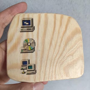 May include: A rectangular wooden coaster with rounded corners featuring three printed pixel art icons. The top icon shows a computer monitor, the middle icon depicts a compact disc, and the bottom icon displays a computer tower and monitor setup.