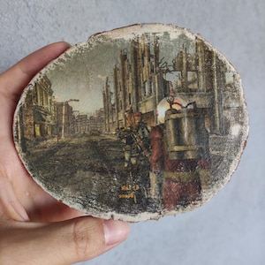 Peut inclure: Une tranche de bois circulaire présentant une illustration imprimée d'une rue de ville post-apocalyptique avec des bâtiments en ruine. Un personnage en costume rouge avec un grand sac à dos métallique est au premier plan. Le texte "Fallout" est imprimé en jaune.