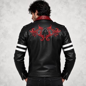 Puede incluir: Una chaqueta de cuero negra con cuello rojo oscuro, dos rayas horizontales blancas en cada manga y un diseño tribal rojo bordado simétricamente en la parte superior de la espalda. La chaqueta incluye charreteras y puños con cremallera.