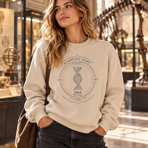 Op de afbeelding: Een beige sweater met ronde hals en een zwarte grafische print van een DNA-dubbelhelix in een cirkelvormig kader. De tekst luidt Scientific Icons No. 02, DNA Double Helix en Discovered 1953, vergezeld door kleine wetenschappelijke symbolen.