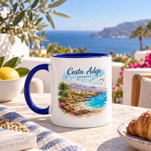 May include: A white ceramic mug with a dark blue handle and interior rim. The front features a colorful watercolor-style illustration of a coastal landscape with palm trees, a beach, and mountains, with the text Costa Adeje TENERIFFA above.