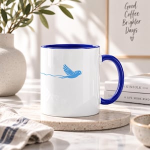 Könnte beinhalten: Ein weißer Keramikbecher mit tiefblauem Innenraum und passendem blauem Henkel. Die Außenseite zeigt eine minimalistische hellblaue Grafik eines kleinen Vogels im Flug, der eine dünne, wellenförmige Linie hinter sich herzieht.