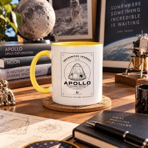 Könnte beinhalten: Ein weißer Keramikbecher mit leuchtend gelbem Innenraum und Henkel. Auf der Vorderseite befindet sich eine schwarze Strichzeichnung eines Apollo-Kommandomoduls mit dem Text Mechanical Legends No. 05, Apollo Command Module, Space Exploration Masterpiece, Est. 1966.
