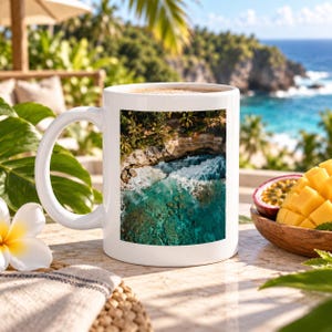 Può includere: Una tazza in ceramica bianca con una fotografia aerea stampata di una costa tropicale con acqua turchese, schiuma marina bianca e scogliere rocciose. La tazza ha un manico curvo ed è posizionata su una superficie chiara vicino a un fiore bianco.