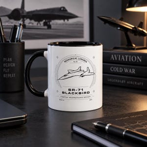 Könnte beinhalten: Ein weißer Keramikbecher mit schwarzem Henkel und schwarzem Innenraum, verziert mit einer schwarzen Strichzeichnung eines SR-71 Blackbird Flugzeugs. Der Text lautet Mechanical Legends, SR-71 Blackbird, Strategic Reconnaissance Aircraft, Mach 3+ und 1964.