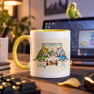 Może przedstawiać: Biały ceramiczny kubek z żółtym uchem i wnętrzem, ozdobiony grafiką przedstawiającą zieloną i niebieską papużkę falistą grające w gry wideo na kontrolerach. Pod ptakami znajduje się napis Eddy i Fly oraz dwie małe butelki i paczka chipsów.