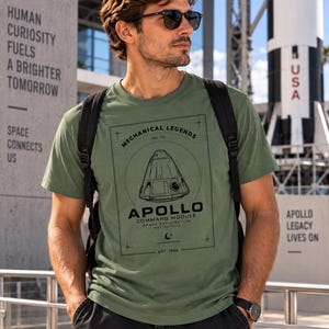 Könnte beinhalten: Ein olivgrünes Kurzarm-T-Shirt mit einem schwarzen Grafikdruck des Apollo-Kommandomoduls. Der Text lautet Mechanical Legends No. 05, Apollo Command Module, Space Exploration Masterpiece und Est. 1966, eingefasst in einem rechteckigen Rahmen.