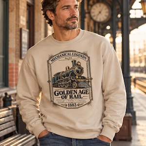 May include: A beige crewneck sweatshirt featuring a graphic print of a vintage steam locomotive. The text reads Mechanical Legends No. 04, Golden Age of Rail, Steam Steel Travel, and 1883. The design is framed in a rectangular border.