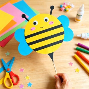 May include: A yellow and black striped bee craft cutout with blue wings, googly eyes, and pink cheeks attached to a wooden stick. The bee features two black antennae and a pointed black stinger at the bottom, creating a cheerful insect design.