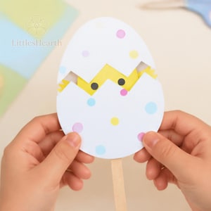 Puede incluir: Un huevo de papel con una cáscara blanca decorada con lunares de colores rosa, amarillo y azul. El huevo está entreabierto para revelar un pollito amarillo con ojos negros en su interior. Un palito de madera está fijado a la base del huevo.