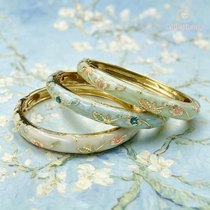 Op de afbeelding: Drie scharnierende armbanden van goudkleurig metaal, elk met een gladde emaille afwerking in tinten wit, lichtgroen en lichtblauw. De armbanden zijn versierd met delicate bloemmotieven in roze, blauw en groen emaille.