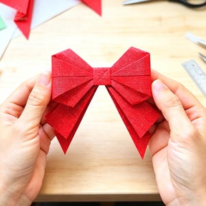 Pode incluir: Um laço de papel vermelho brilhante com uma superfície texturizada, apresentando um nó quadrado central e dois laços plissados simétricos de cada lado, com duas pontas afiadas estendendo-se para baixo a partir da base do laço.